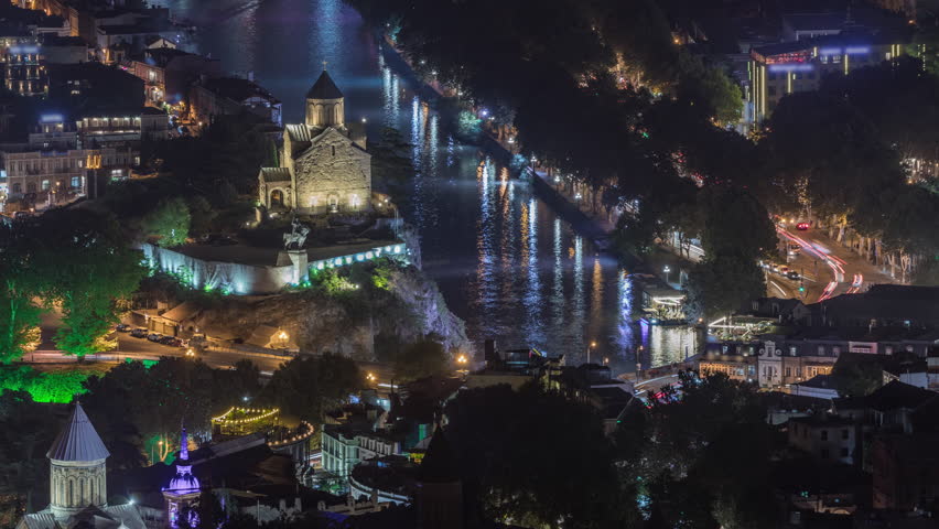 Metekhi Virgin Mary Assumption Church aerial night timelapse and the Statue of King Vakhtang Gorgasali above the Kura River. Traffic on a bridge and illuminated waterfront. Tbilisi, Georgia.