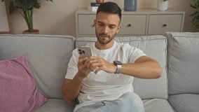 Young hispanic man with a beard in a white shirt sitting on a sofa at home, using a smartphone and checking his smartwatch in the living room. - Powered by Shutterstock - Get 15% off with code: PIKWIZARD15