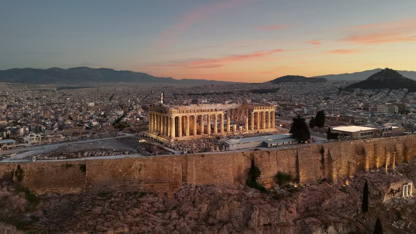 Flying around Parthenon in Athens, Greece at sunrise. Acropolis with city lights and Athens city below. Top international landmark in Greece. Aerial shot, UHD