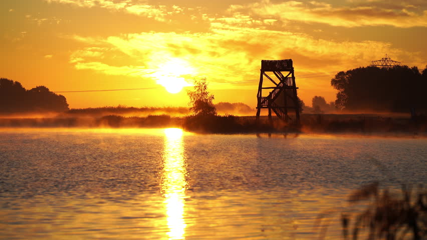 A wooden overlook tower at a lake during a tranquil, foggy sunrise in autumn.