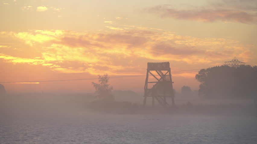 A wooden overlook tower at a lake during a tranquil, foggy sunrise in autumn.