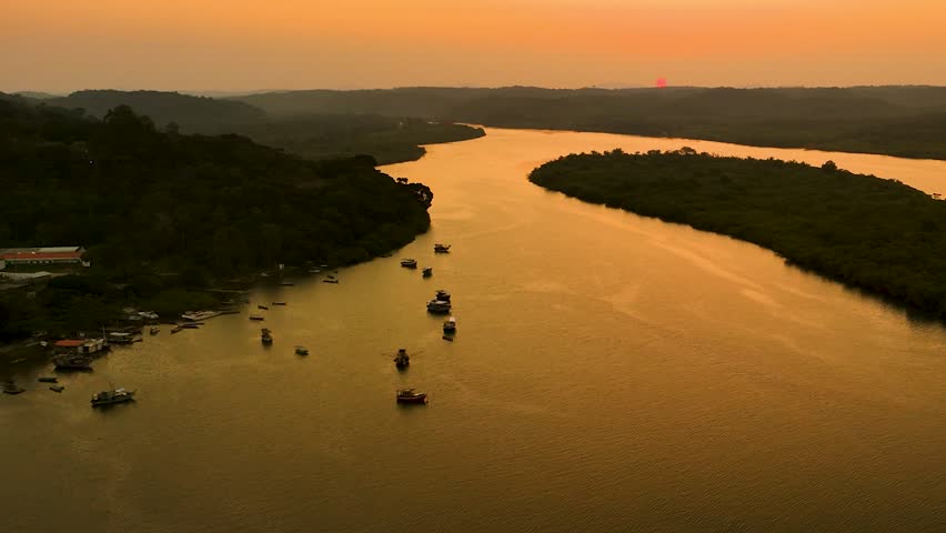 Aerial over a forest river with boats, an island, and a pink sun and orange sunset