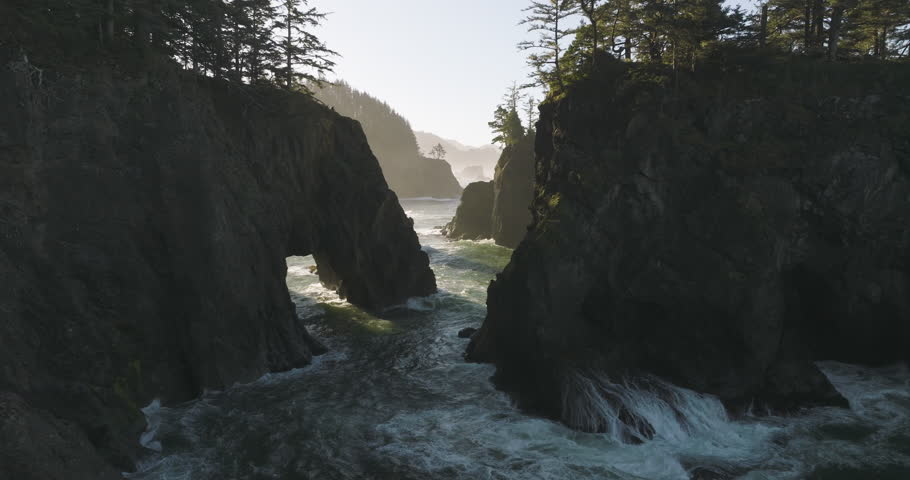 Aerial view of waves crashing through the rocky natural bridges along the Oregon coast, surrounded by forested cliffs and rugged terrain