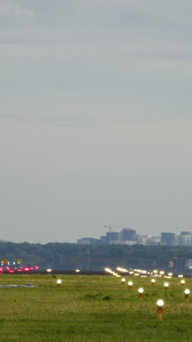 Vertical video of a large airplane touching down on a runway with its landing gear extended, lights lining the airstrip. The background features a soft skyline with no people present