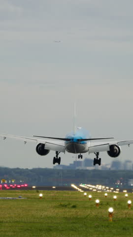 Vertical video of a large airplane touching down on a runway with its landing gear extended, lights lining the airstrip. The background features a soft skyline with no people present