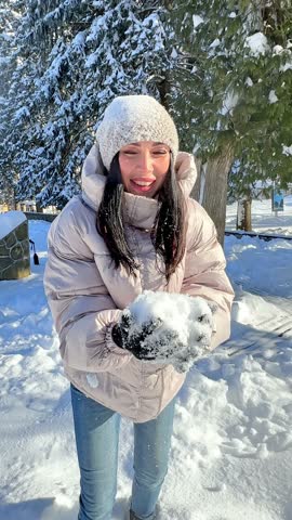 Happy young woman in warm winter clothes holding snow in her hands and blowing on it, snowflakes flying towards the camera. Fun in winter city park. Slow motion vertical footage. Outdoor entertainment