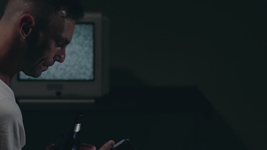 Man in White T-shirt is Sitting Against the Old Silver TV in Dark Room. Drinking Beer and Using His Smartphone. Search for TV Signal. Distortion on the Screen. Back VIew, Medium Close-Up, Film Grain