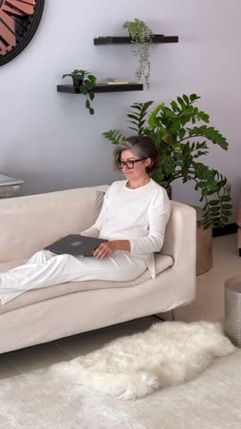 Focused gray-haired woman seated on beige sofa, wearing white clothes, smiling while typing laptop, greenery behind her, relaxed cozy vibe, modern home, soft lighting.