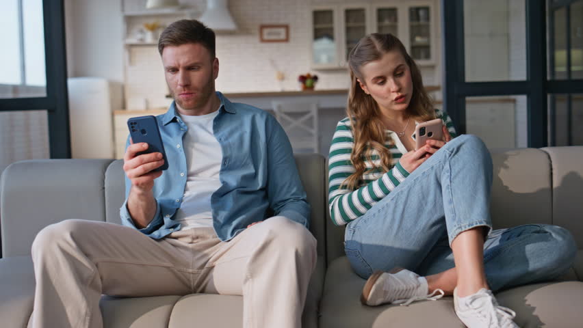 Two people using smartphones sitting soft couch. Focused married couple typing mobile phones feeling bored in apartment. Soulmates with cellphones turning heads looking each other with smile indoor 