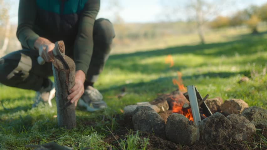 A man chops wood with an axe by a campfire in the wilderness, surrounded by greenery, focusing on survival techniques and the necessity of fire preparation for camping.