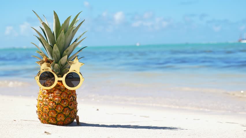 Funny pineapple face in funky sun glasses on caribbean seashore. Exotic fruit on beach close to sea. Summer holidays