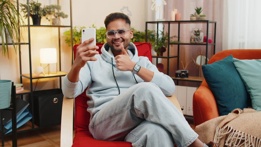 Happy Indian man blogger taking selfie on smartphone, communicating video call online with subscribers, recording stories for social media. Guy at modern home apartment living room sitting on chair