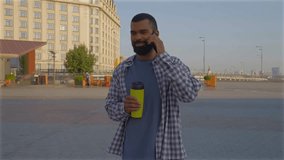 Positive smiling handsome bearded African American man in trendy casual outfit holding travel cup, friendly chatting with friend using mobile phone while standing on city street . - Powered by Shutterstock - Get 15% off with code: PIKWIZARD15