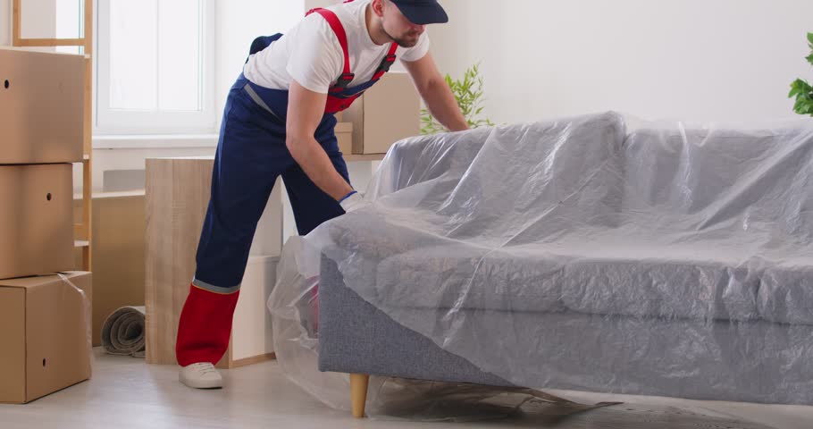 Moving service employee worker moving heavy sofa in living room full of cardboard boxes. Man worker from delivery service placing furniture in new apartment. Relocation concept. 4k video