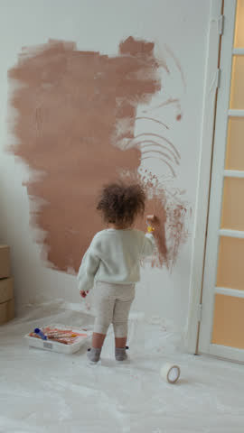 Vertical fu shot of little curly-haired baby girl painting wall brown with mini roller during room renovation in new apartment