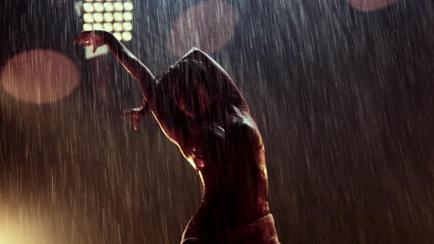 A dancer showcases dynamic hand movements in heavy rain, with vibrant orange lighting illuminating the scene and enhancing the energy of her performance