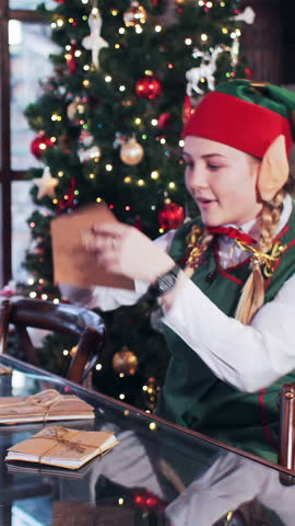 An elf girl sits in a room with a Christmas tree and looks through the letters to the children of Santa Claus. Vertical video.