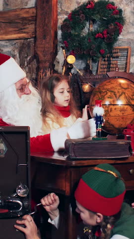 Santa Claus and children are exploring the country on a retro globe. Christmas elves sitting on the floor set up an old gramophone. Vertical video.