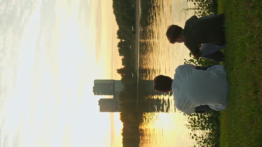 Father and son are seated on the grass in a public park, facing the water, taking in the city skyline as the sun