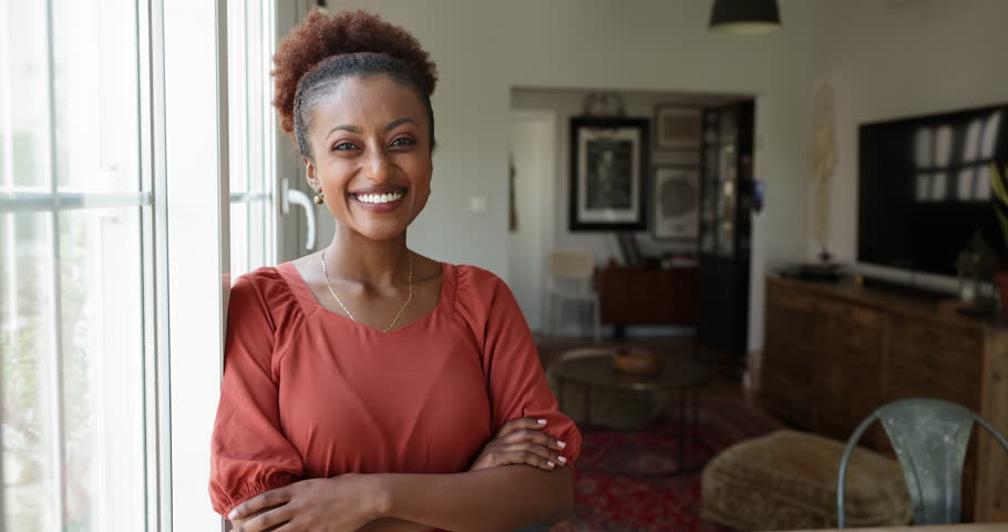 Curly haired African female stand near window with arms-crossed looks at camera posing in cozy living room. Satisfied homeowner, housewife, tenant or renter. Independent Ethiopian woman home portrait