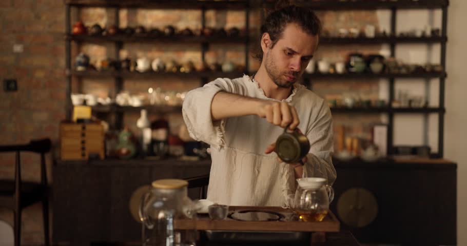 Confident and calm guy with curly hair and stubble pours tea from a ceramic teapot during a desperate ceremony in a tea cafe