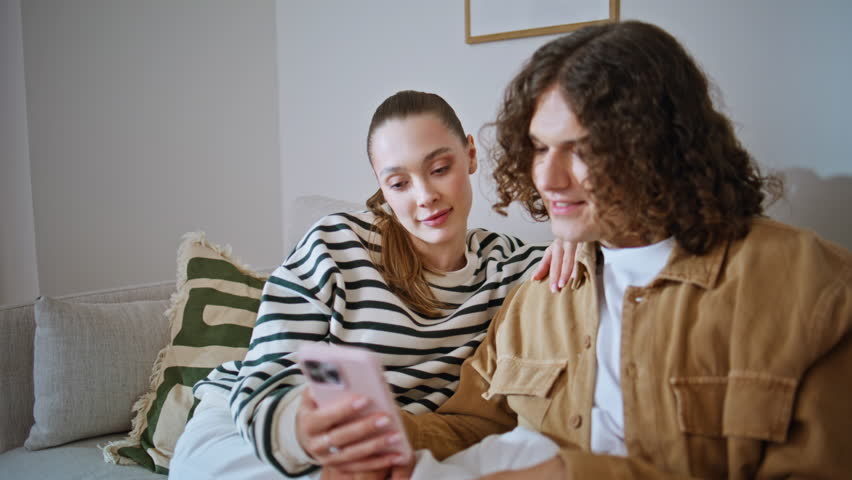 Romantic couple sharing smartphone watching social media content at comfortable sofa closeup. Relaxed woman caressing curly man hair looking together at cellphone screen. Loving spouses feeling happy.