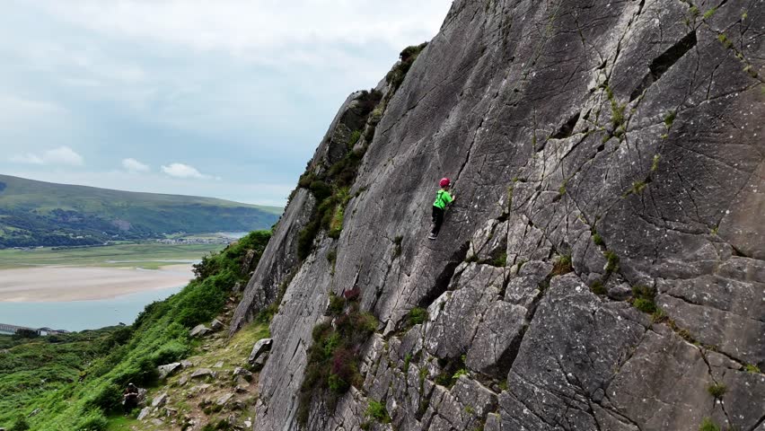 A young climber ascending the dramatic Barmouth slabs in North Wales, with stunning views of hills and coastlines below.