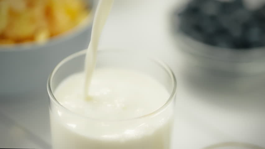 Fresh milk is being poured into a clear glass, surrounded by a vibrant breakfast of fruits and cereals, creating a healthy morning atmosphere in a sunny kitchen