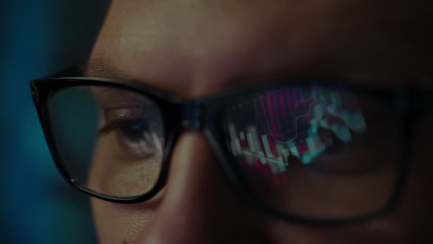 A person with glasses is intently watching a computer screen illuminated by colorful lights. The close-up emphasizes their focused expression and the glow reflecting in their eyes
