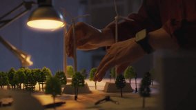 A person meticulously arranges miniature trees and wind turbines in a workspace illuminated by warm light, showcasing attention to detail in landscape design during the evening - Powered by Shutterstock - Get 15% off with code: PIKWIZARD15