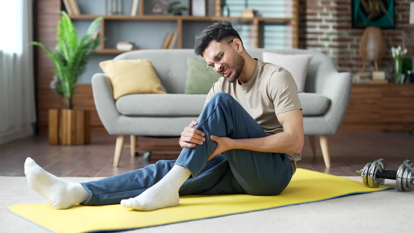 Young adult man suffers from leg pain, calf muscle spasm during a gymnastics workout sitting on the floor on a mat in the living room at home. Male massaging leg calf muscle cramps tendon at calves