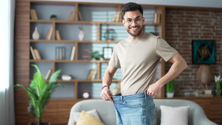 Happy handsome man feels joy after losing weight. Smiling satisfied adult bearded male tries on his big pants jeans, which he wore before, standing in living room at home looking at the camera