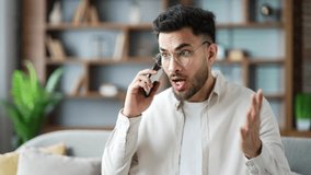 Angry excited man talking arguing on smartphone sitting on sofa in apartment at home. Frustrated young man shouting on phone call, expressing stress and anger emotions in modern living room. Close up - Powered by Shutterstock - Get 15% off with code: PIKWIZARD15