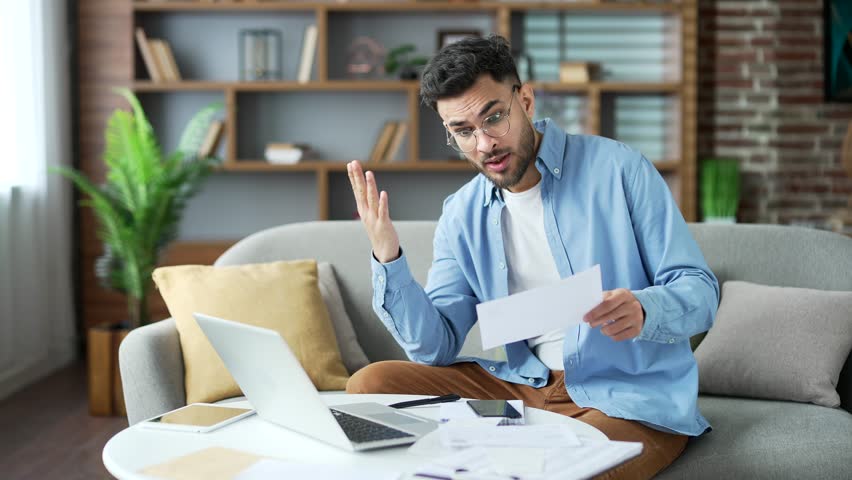 Frustrated young adult man looking at large utility bills sitting on sofa at home office. Upset bearded male holding a paycheck, thinking about problems in the household budget and financial expenses - Powered by Shutterstock - Get 15% off with code: PIKWIZARD15