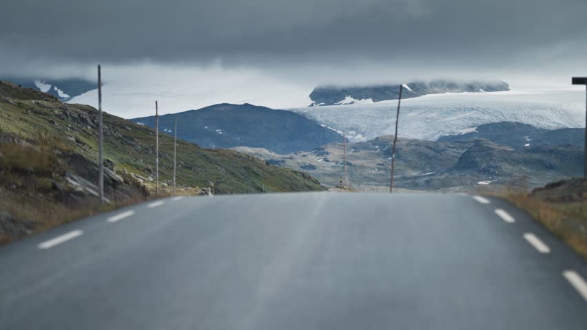 The road curves through a rugged mountain scene, surrounded by boulders and leading toward distant mountain peaks and glaciers.