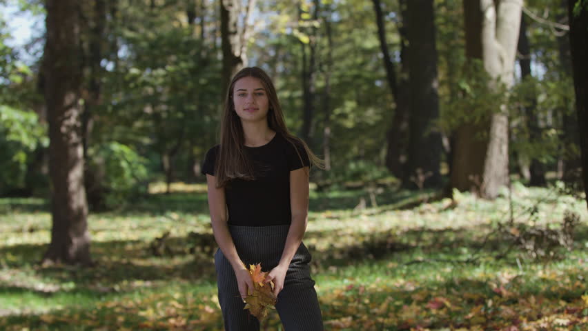 girl is covering her face and waving a maple tree branch with yellow leaves in autumn park. Slow motion
