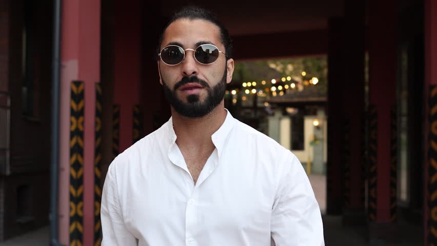 A dynamic image of a waist-length handsome man with a beard, wearing a white shirt, sunglasses and black glasses. Slow motion.
