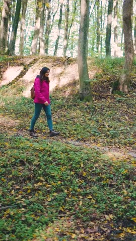 Attractive young woman in a pink jacket, jeans, and backpack walking through an autumn forest, crossing a wooden footbridge, slow motion