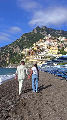 A couple enjoys a romantic walk along the pebbled shoreline of the Positano Amalfi coast, hand in hand, with colorful houses adorning the hillside under a bright blue sky in Italy