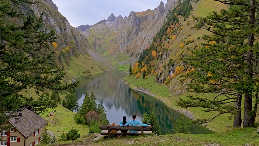 Two friends enjoy a peaceful moment on a bench overlooking a stunning lake, framed by vibrant autumn foliage and majestic mountains in the Swiss Alps. Falensee lake, Appenzell Innerrhoden, Switzerland