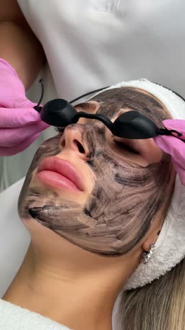 Beautician doing hardware carbon peeling to a young beautiful client in a headdress in goggles lying on a sofa in a beauty salon. Facial cleansing concept. Cosmetology services