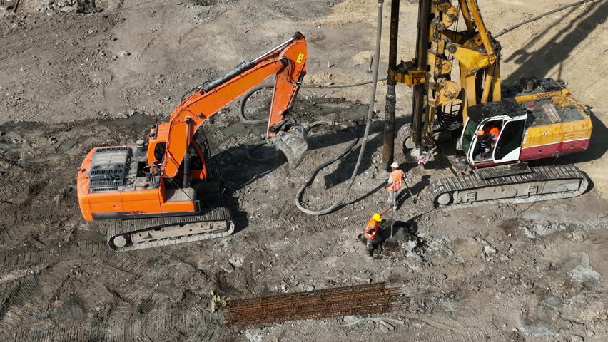 Construction workers operating heavy equipment at a job site. An aerial view of a construction site with workers operating large excavators and drilling machinery.