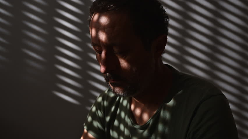 Regret. Regretful mature adult male sitting alone in dark room. 4K with selective focus.