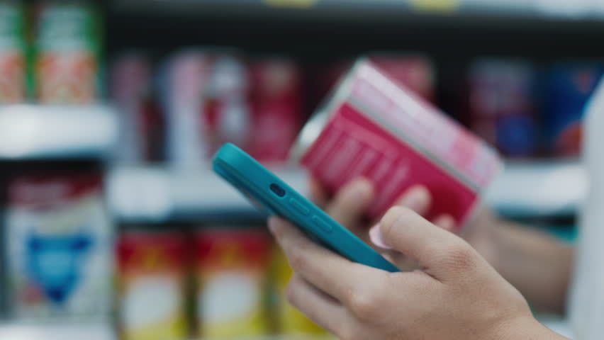 Delicate Hands Of Woman Holding Can While Reading Ingredients On Phone. Young Female Uses Calorie Calculator App On Her Smartphone To Track Nutritional Information. Shopping Concept