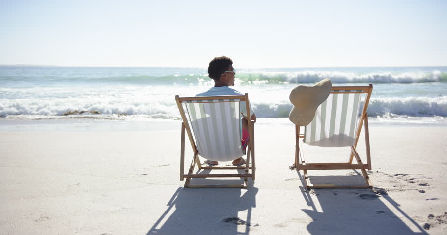 A young biracial man relaxes on beach chair facing ocean, copy space. An empty chair beside him suggests a companion might return soon, unaltered, slow motion