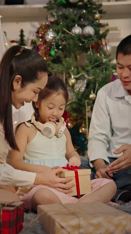 Happy Asian family exchanging presents on Christmas morning celebrating winter holiday Together. Cute little girl receiving gifts box from her father and mother sharing New Year celebration moment.
