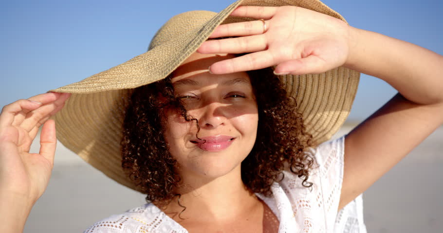 A young biracial woman with curly hair smiles on a sunny beach, shielding her eyes with her hand. She wears a wide-brimmed hat and a white outfit, radiating joy in the daylight.