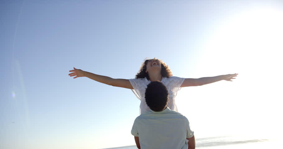Biracial couple embraces on a sunny beach, with clear blue skies overhead, with copy space. The woman's curly hair catches the light as they share a joyful moment by the sea, slow motion.