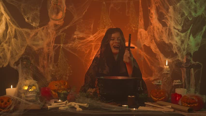 A witch in black clothing conjures up a scary kitchen with pumpkins and human skull and bones. A young woman picks up and turns over a cross for a ritual and laughs eerily. Halloween Concept.