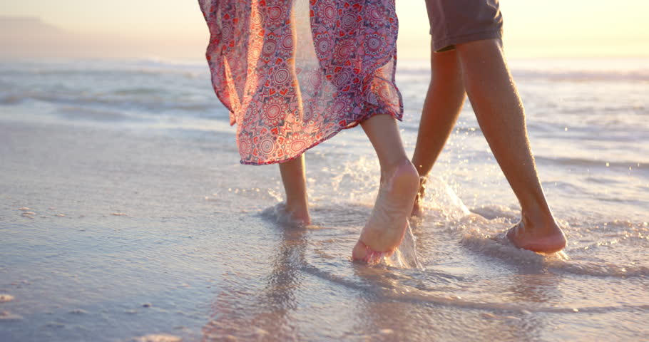 Biracial couple walks barefoot on the beach at sunset, woman wearing a patterned dress, with copy space. Gentle waves and the warm glow of the evening sun create a romantic setting, slow motion.